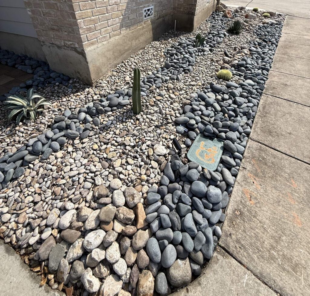 Oakwell Farms Xeriscape & Landscape Lighting Mini dry creek bed of black Mexican beach pebble running through tan river rock beside a sidewalk, with a fence post cactus and variegated agave