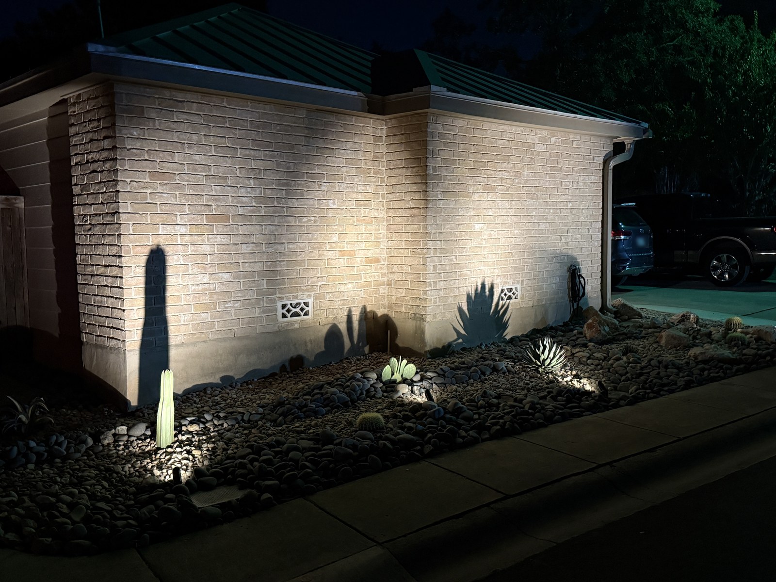 Oakwell Farms Xeriscape & Landscape Lighting At night: low-voltage uplights casting cactus and agave shadows on a brick wall above a river rock bed