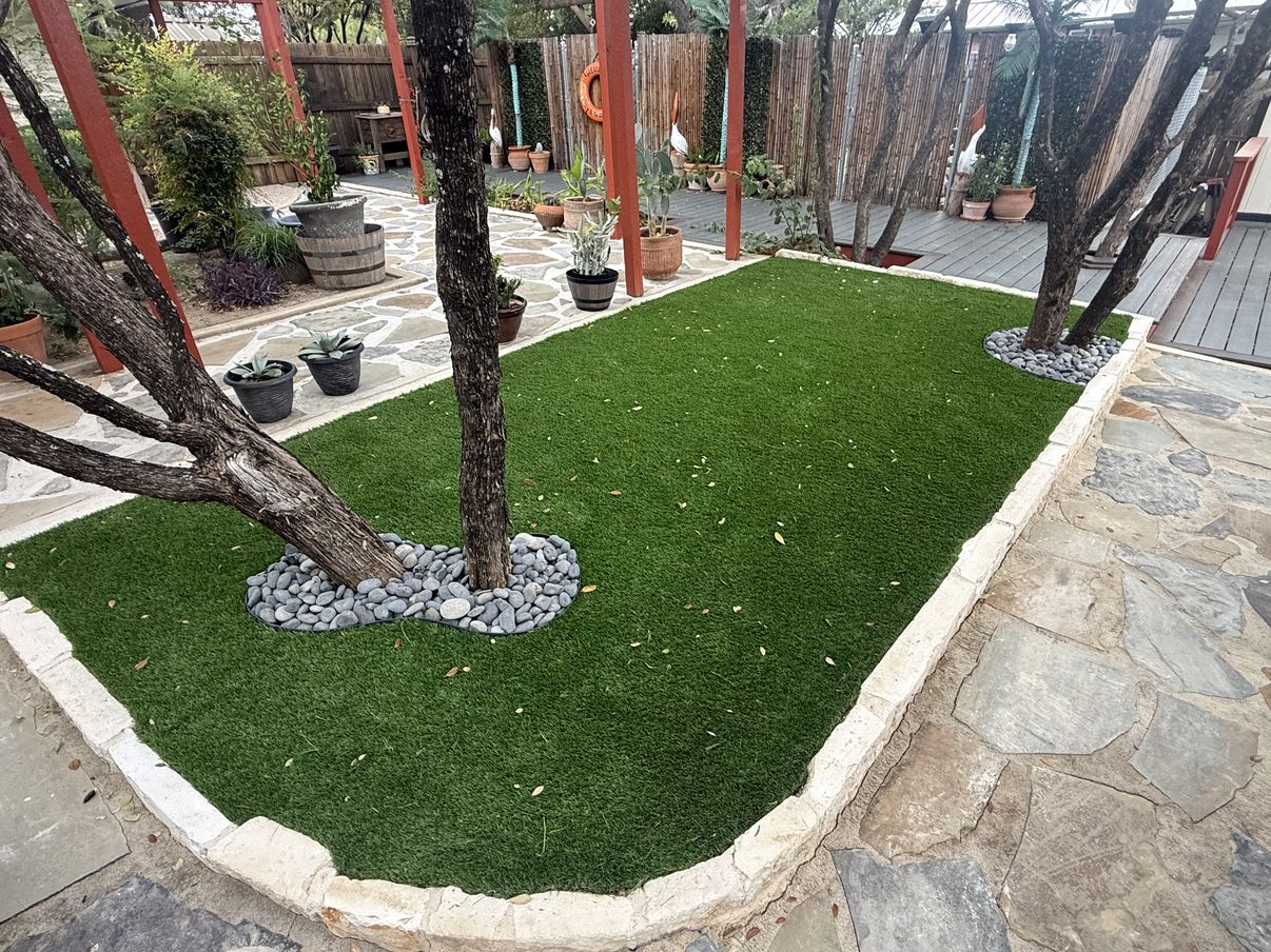 Same backyard after: artificial turf with a limestone border and river-rock rings around the trees