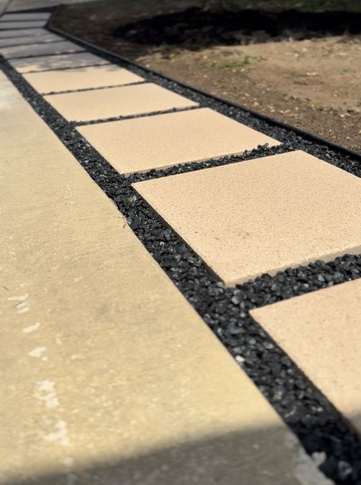 Large tan stepping-stone pavers set in black rock with steel edging beside a driveway