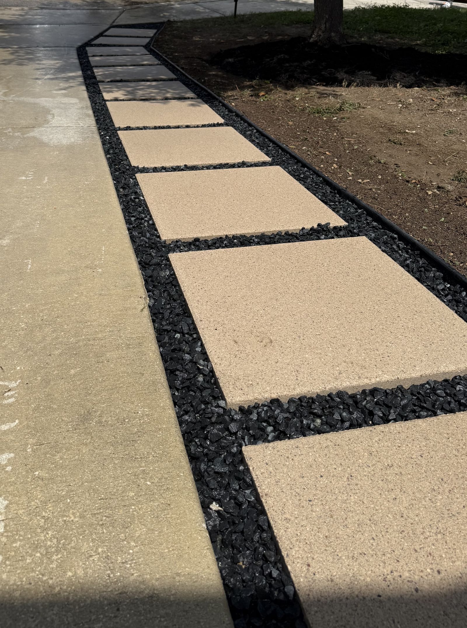Large tan stepping-stone pavers set in black rock with steel edging beside a driveway