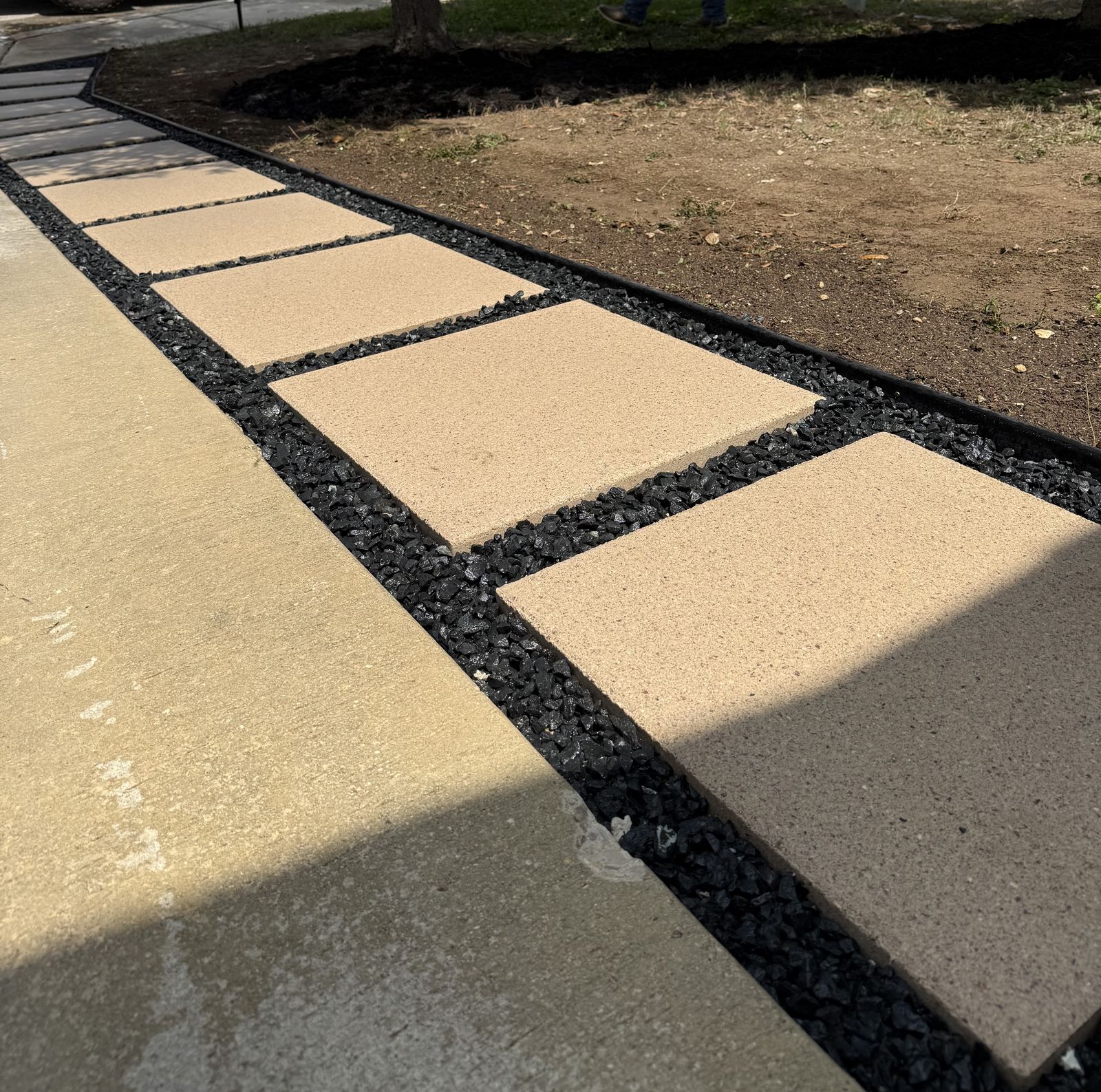 Large tan stepping-stone pavers set in black rock with steel edging beside a driveway